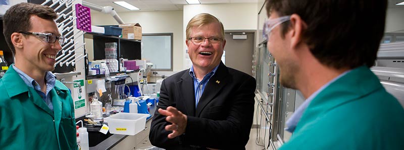 Blond man in a suit talks with two researchers in a lab